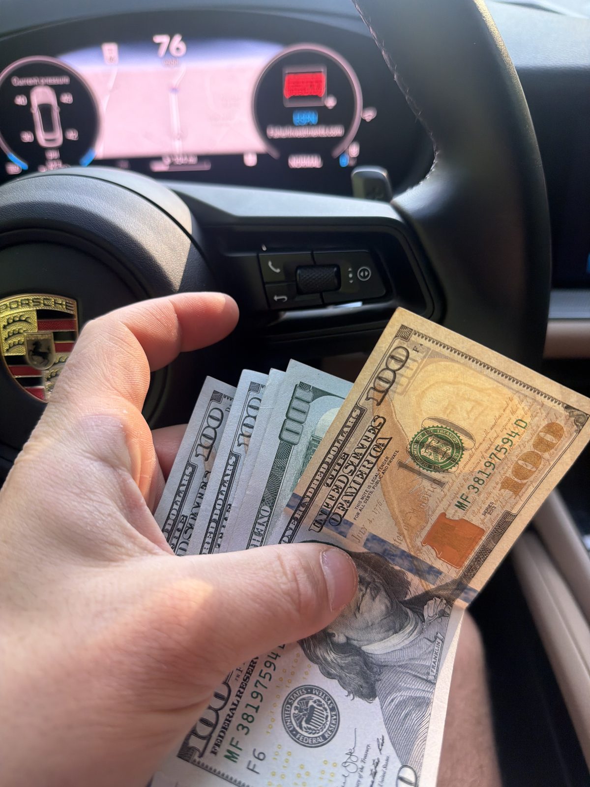 Hand holding hundred dollar bills in front of a Porsche steering wheel